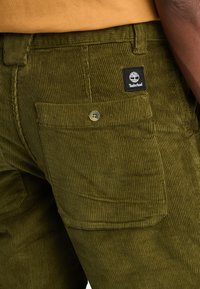 I pantaloni di corduroy verde oliva presentano una superficie testurizzata, una tasca cargo laterale e una toppa del logo Timberland nera sopra la tasca posteriore.