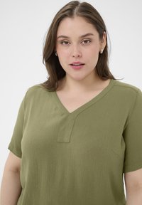 Haut manches courtes vert olive avec un col en V et un tissu texturé. Il comprend une poche discrète sur la poitrine et une coupe décontractée.