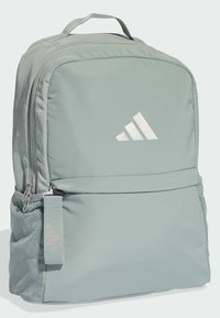 Mochila de color gris claro con una textura suave y lisa. Presenta un prominente logo de marca blanco, un bolsillo frontal con cremallera y un bolsillo lateral de malla.