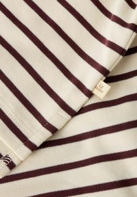 Raidallinen puuvillakangas, jossa on kermaista ja burgundia, tasaiset vaaka-raidat, pehmeä rakenne ja pieni näkyvä etiketti reunassa.