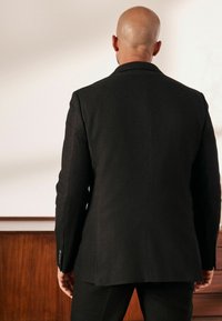 Blazer negro a medida con un diseño estructurado, que presenta solapas de muesca, una abertura central y puños abotonados. Textura de tela suave.