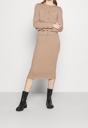 Nainen, joka käyttää beigeä ribbineulottua napillista neuletakkia ja vastaavaa midihametta, yhdistettynä mustiin nahkakenkiin nilkassa.