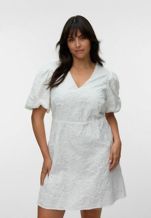 Femeie cu păr închis la culoare poartă o rochie albă cu mâneci scurte și broderie florală, stând în fața unui fundal alb simplu.