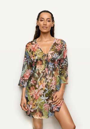 Femme portant une robe transparente à motif floral, manches 3/4, nouée sur le devant avec une fente dévoilant sa cuisse, se tenant devant un fond uni.