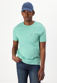 Mint green short-sleeve t-shirt with a chest pocket. Paired with blue denim jeans and a blue cap featuring a logo accent.