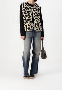 Person som bär en leopardmönstrad väst över en svart långärmad skjorta, bleknade blå jeans, svarta sneakers och håller en brun handväska vid sidan.