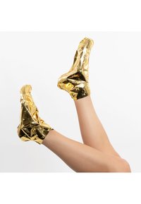 Botines de foiledorado con patrones de estrellas, textura brillante y diseño ajustado al tobillo, mostrados con las piernas levantadas sobre un fondo blanco.