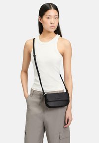 Czarna skórzana torebka na ramię o eleganckim designie, z klapą typu fold-over i regulowanym paskiem, noszona na białym topie na ramiączkach z casualowymi szarymi spodniami.