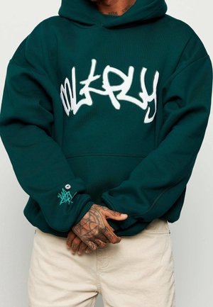 Personne portant un sweat à capuche vert foncé avec un texte blanc audacieux de style graffiti, un pantalon beige et les mains tatouées jointes devant.