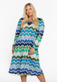Langärmliges Kleid im mehrfarbigen Zickzackmuster mit grünen, blauen und beigen Farbtönen, runden Ausschnitt und dekorativem Ringakzent.