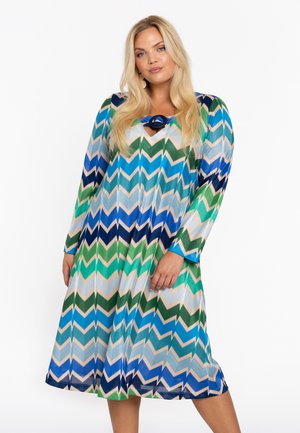 Robe à manches longues avec un motif en zigzag multicolore présentant des tons de vert, bleu et beige, avec un col rond et un accent décoratif en forme d'anneau.