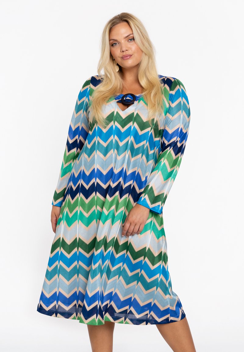 Langärmliges Kleid im mehrfarbigen Zickzackmuster mit grünen, blauen und beigen Farbtönen, runden Ausschnitt und dekorativem Ringakzent.
