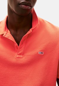 Camisa polo en coral brillante, con un tejido texturizado, cuello corto y un pequeño logotipo en contraste en el pecho.