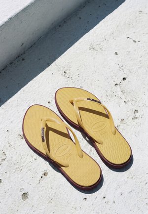Tongs jaunes avec semelles bordeaux reposant sur un béton blanc texturé sous un soleil éclatant.