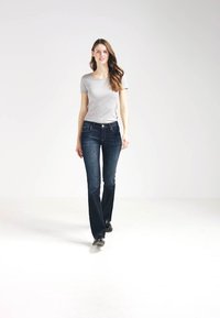 Tummansiniset leveälahkeiset farkut, joissa on viisi taskua ja vaaleat tikkaukset, yhdistettynä kätevään harmaaseen T-paita. Malli kävelee valkoista taustaa vasten.