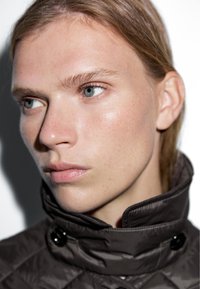 Hnědá prošívaná bunda s vysokým límcem a černými knoflíky, zdůrazňující hladkou pleť a modré oči.