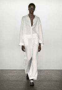 Chemise en lin blanc avec une coupe ample, associée à un pantalon large assorti. Les accessoires comprennent des lunettes de soleil noires et des sandales plates.