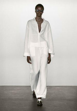 Chemise en lin blanc avec une coupe ample, associée à un pantalon large assorti. Les accessoires comprennent des lunettes de soleil noires et des sandales plates.