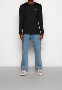 Svart långärmad t-shirt med en vit logotyp på bröstet, kombinerad med ljusblå, löst sittande jeans och vita sneakers med röda detaljer.