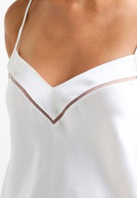 White satin top featuring a V-neckline with a transparent mesh inset, thin straps, and smooth texture. No visible patterns or hardware details.