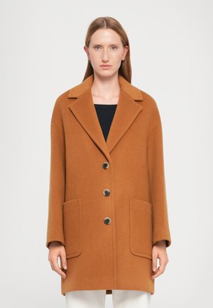 Femme portant un manteau oversize de couleur camel avec un large col, trois grands boutons et deux poches plaquées à l'avant, se tenant contre un fond blanc.