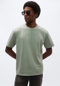 Svetlozelené bavlnené tričko s okrúhlym výstrihom, krátkymi rukávmi a hladkým povrchom, kombinované s čiernymi obdĺžnikovými slnečnými okuliarmi.