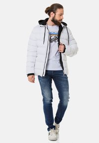 Weißer Pufferjacke mit schwarzen Akzenten und Kapuze, mit gestepptem Design. Getragen über einem weißen Grafik-T-Shirt und blauen Jeans, mit weißen Sneakers.