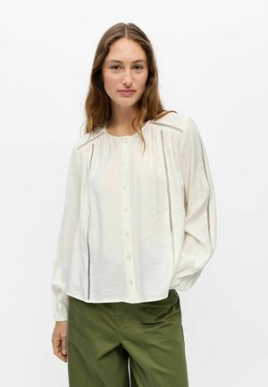 Femme aux longs cheveux bruns portant un chemisier blanc boutonné avec des découpes verticales et un pantalon vert sur un fond blanc uni.