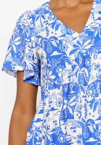 Blauw en wit gestippelde jurk met korte mouwen, een v-hals en grafische ontwerpen van palmbomen, zonnen en landschappen. Lichtgewicht stof.