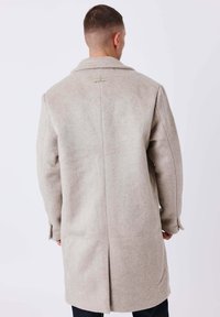 Cappotto beige in misto lana con collo a revers, spacco posteriore e tasche laterali, caratterizzato da una texture sottile e linee pulite.