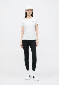 Wit aangesloten T-shirt met een klein logo, gecombineerd met zwarte legging en witte sneakers. Accessoires zijn onder andere een witte pet en witte sokken.