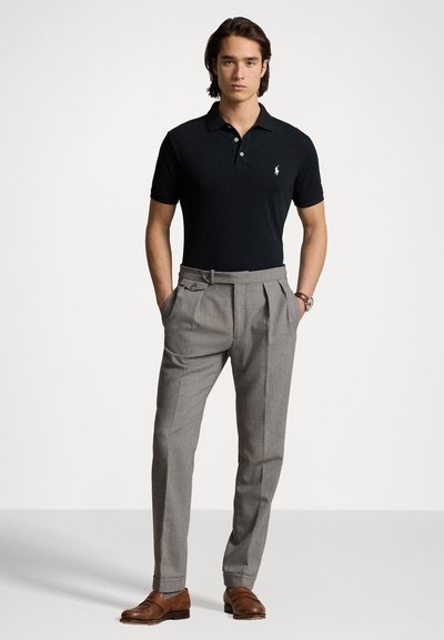 Sort polo shirt med krave og tre knapper, parret med grå plisserede bukser. Brune lædersko med struktureret overflade.