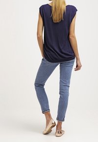 Top blu navy largo con maniche a leggenda, abbinato a jeans skinny blu chiaro e ballerine dorate metalliche, visto da dietro.