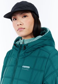 Jong persoon met lang donker haar, gekleed in een zwarte pet en een teal gewatteerde hooded jack met het "PROTEST" logo op de borst.