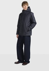 Voděodolná námořnická bunda s kapucí, patentkami a dvěma postranními kapsami, spojená s kalhotami širokého střihu z tmavého denimu a hnědými koženými botami.