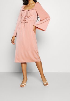 Różowa sukienka w odcieniu pudrowego różu z kwadratowym dekoltem, marszczonym stanem i długimi rozkloszowanymi rękawami. Gładki materiał i wiązana talia. Noszona z białymi szpilkami.