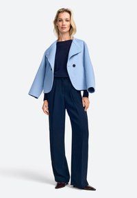 Giacca corta azzurro chiaro con un ampio collare e due bottoni neri. Abbinata a pantaloni a gamba larga blu navy e scarpe scure, su uno sfondo semplice.