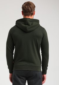 Sudadera de color verde oscuro con capucha ajustable, mangas largas y puños acanalados. Textura suave con una etiqueta de marca visible en el lado inferior izquierdo.