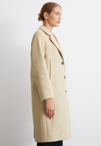 Beige Wollmantel mit Schalkragen, knöpfbarem Einreißverschluss, seitlichen Taschen und lockerer Passform, seitliche Ansicht vor einem neutralen Hintergrund präsentiert.