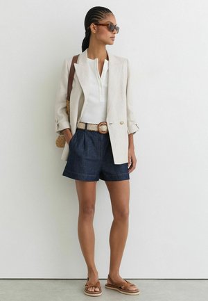 Femme aux cheveux tressés porte des lunettes de soleil, un blazer beige, une chemise blanche, un short en jean avec une ceinture tissée, portant un sac tissé et des sandales marron.