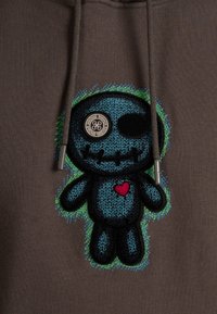 Felpa marrone con un grande personaggio cartoon ricamato in blu con un cuore, dettagli neri e un design testurizzato sulla parte anteriore.