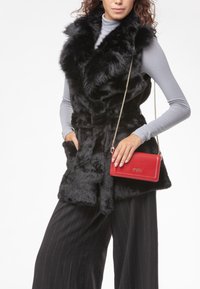 Completo in ecopelliccia nera con un ampio colletto, abbinato a una borsa a tracolla in pelle rossa. Il modello indossa una maglia grigia a maniche lunghe e pantaloni con motivo.