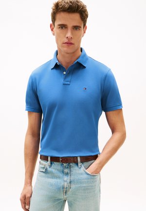 Fiatal férfi kék pólóingben kis logóval, világoskék farmerral és barna övvel, egyik kezével a zsebében áll.