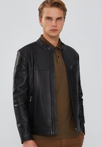 Zwarte leren jas met een strak ontwerp, voorzien van een staande kraag, ritszakken en een soepele textuur. Draagt over een bruin polo-shirt.