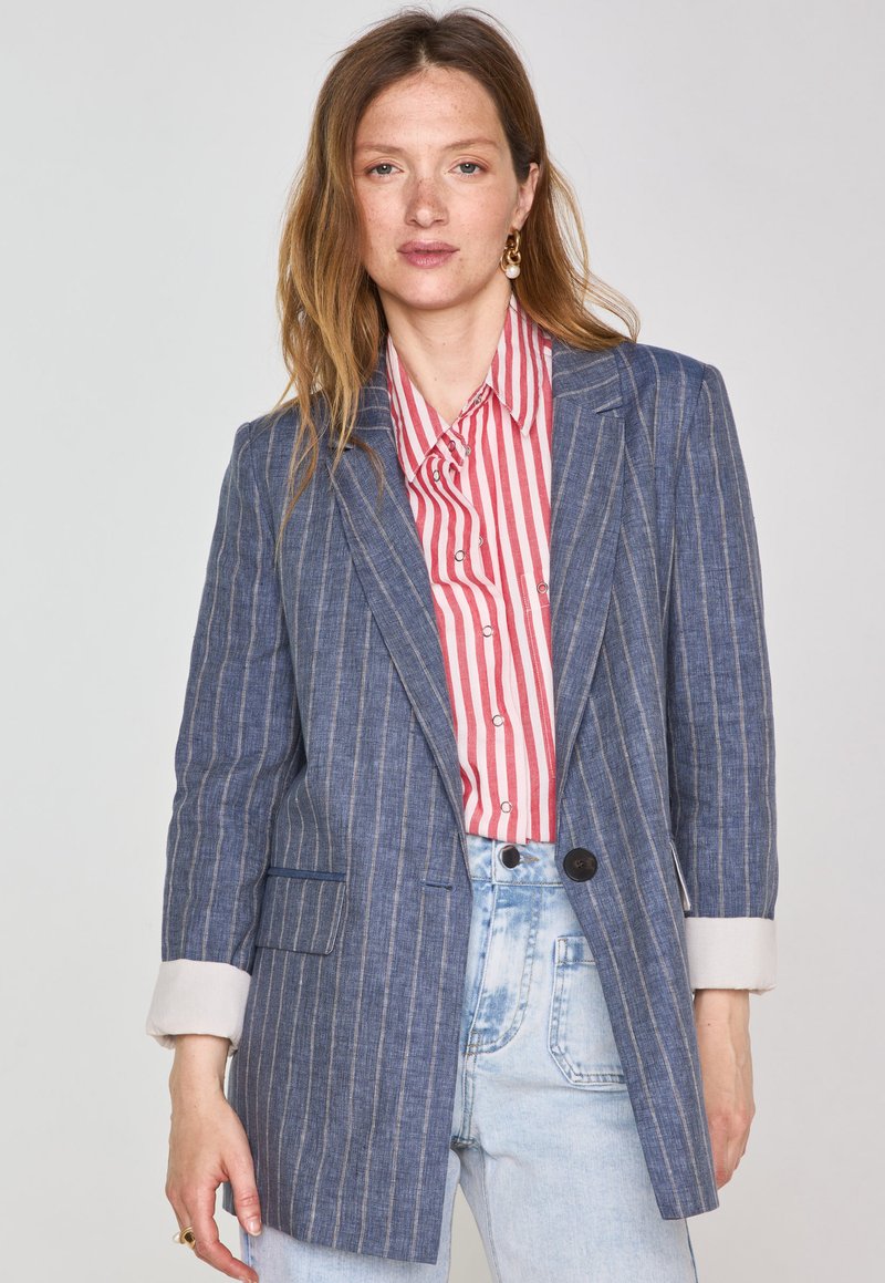 Blazer bleu à rayures fabriqué en tissu léger avec une fermeture à un bouton, associé à une chemise rayée rouge et blanche.