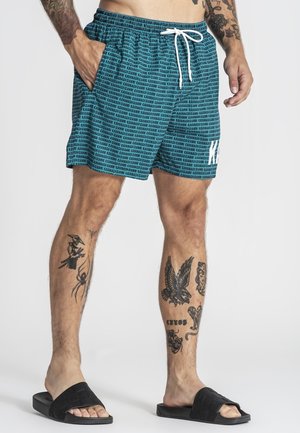 Muž v tyrkysových krátkych nohaviciach s bielymi šnúrkami, čiernych slidových sandáloch a s viacerými tetovaniami na nohách a rukách, stojaci s rukami v kapsách.