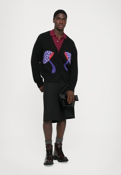 Bărbat îmbrăcat cu un cardigan negru cu modele de ciuperci purpurii, tricou polo bordeaux, pantaloni scurți negri până la genunchi, cizme maronii, șosete gri, ținând în mână o poșetă neagră.