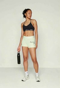 Woman wearing black sports bra and light shorts holding black water bottle, standing with legs apart against plain background.