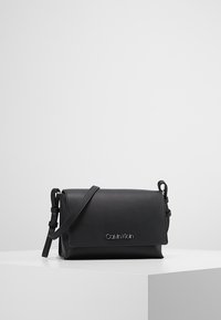 Černá kožená crossbody taška s designem klapky, stříbrným logem, nastavitelným popruhem, hladkým povrchem a obdélníkovým tvarem.