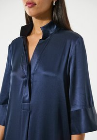 Camicetta di raso blu navy con colletto alto e maniche corte a campana. Presenta una parte di pattina frontale e una consistenza liscia con una leggera lucentezza.